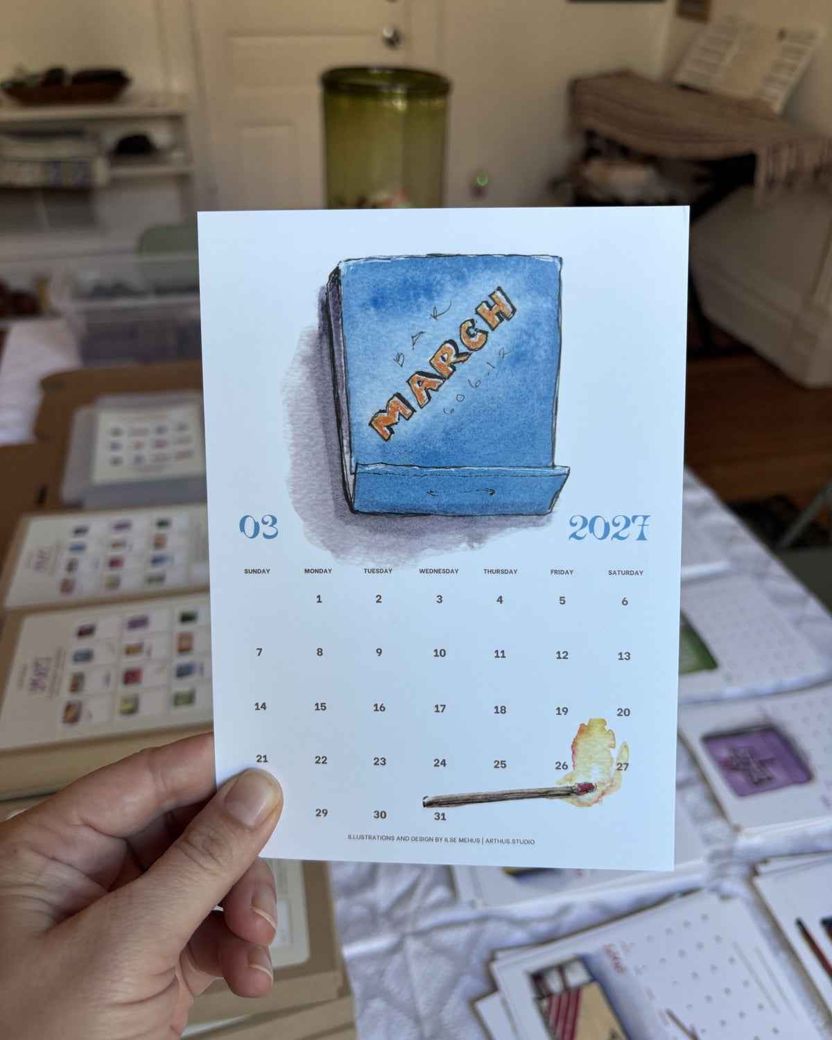 march 2027 postcard from the arthus calendar held in hand, blue watercolor matchbook inspired by bar primi