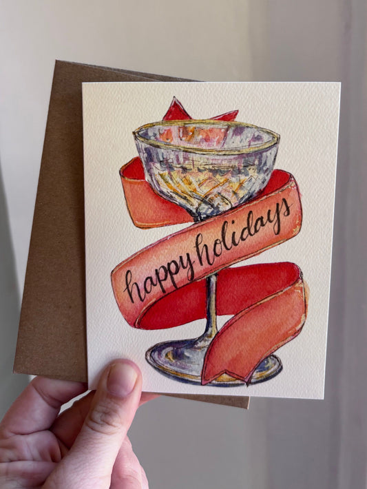 happy holidays champagne watercolor greeting card by arthus, held in hand with a kraft envelope